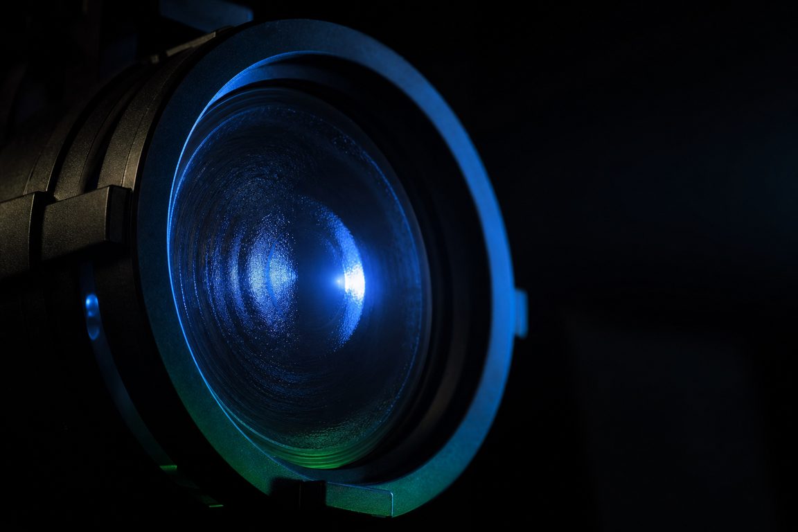 A stage spotlight lens seen head-on in darkness, flare across the glass